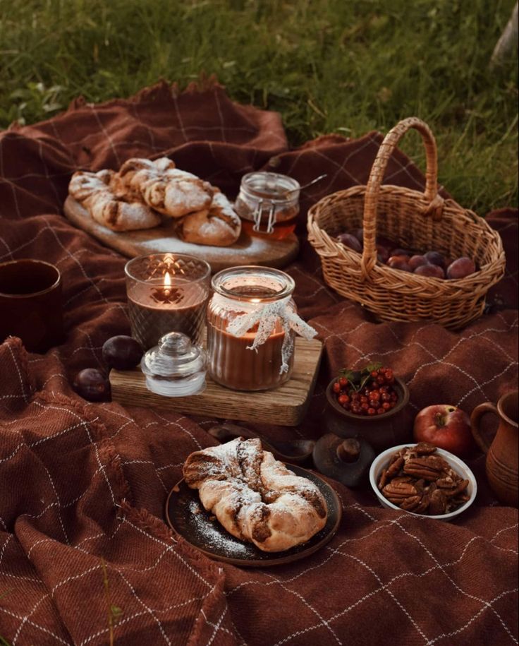 Cozy autumn picnic setup with pastries, plums, nuts, candles, and drinks on a checkered blanket outdoors.