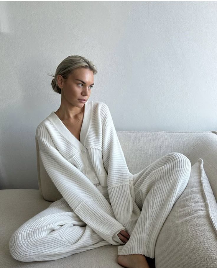 Woman in cozy white loungewear sitting on a cream sofa, gazing thoughtfully to the side in a minimalist setting.