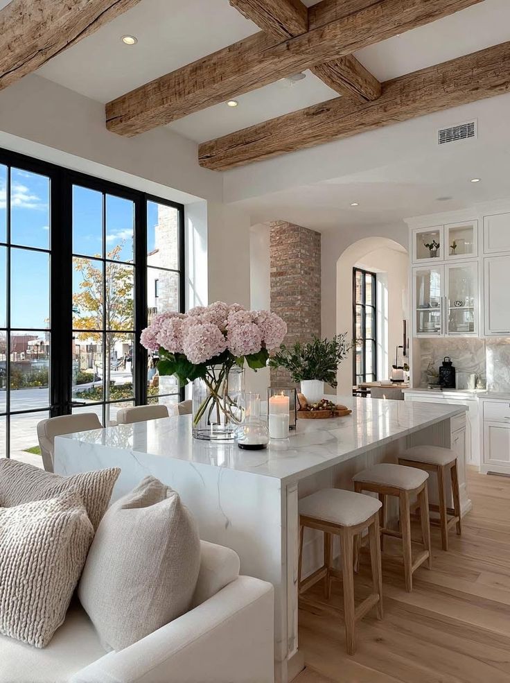 Modern kitchen with wooden beams, marble island, and large windows, featuring pink flowers and candles for decor.