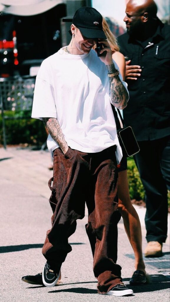 Young man in casual clothing, smiling on phone while walking outdoors, wearing a white t-shirt and brown pants.
