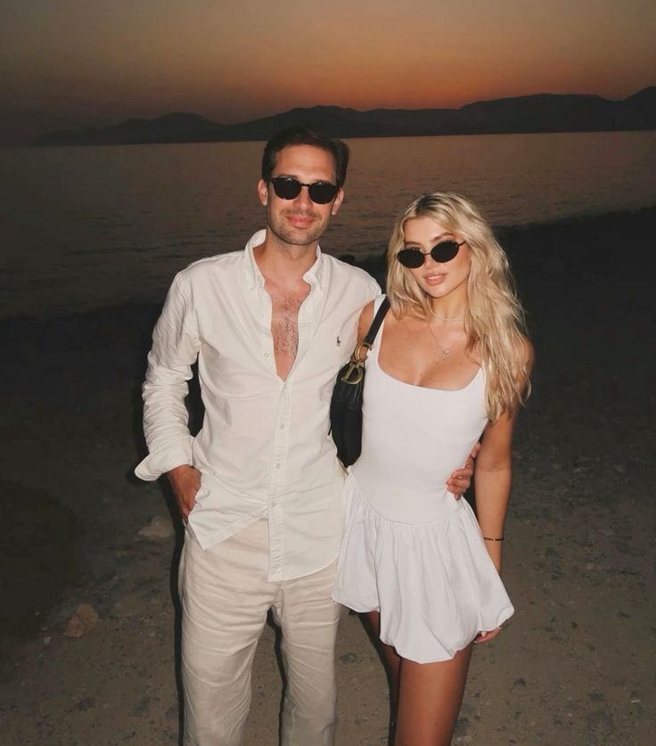 Couple in white outfits posing by a serene sunset beach backdrop, both wearing stylish sunglasses.