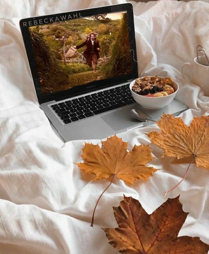 Laptop on bed with fall leaves, streaming a movie. Bowl of cereal and cozy ambience complete the autumn setup.