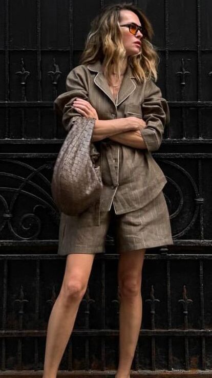 Woman in chic brown outfit with shorts and jacket, holding a large bag, wearing stylish sunglasses and sandals.