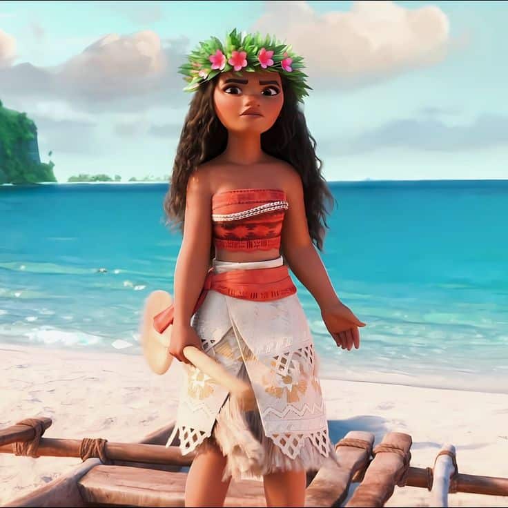 Animated character in traditional attire stands by a beach, wearing a floral crown. Ocean and clouds in the background.