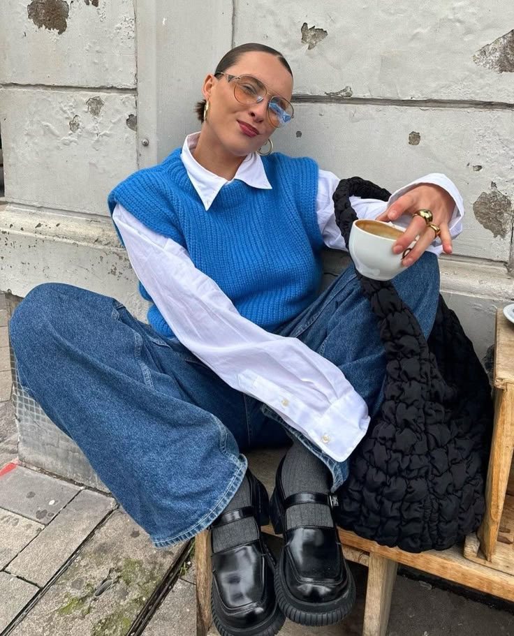 Woman in blue sweater vest and jeans enjoys coffee seated outdoors with a stylish black bag.