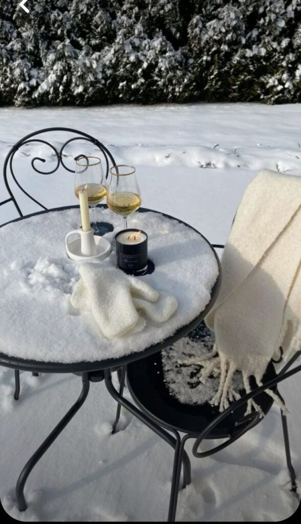 Snowy outdoor table with wine glasses, candle, and gloves on chairs, creating a cozy winter setting.