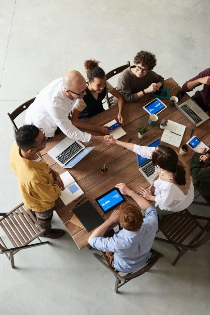 Colleagues collaborating around a table with laptops and tablets, shaking hands in a modern workspace.