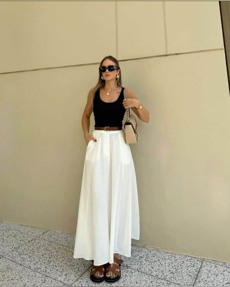 Woman in stylish summer outfit with black top, white skirt, sunglasses, and woven bag in urban setting.