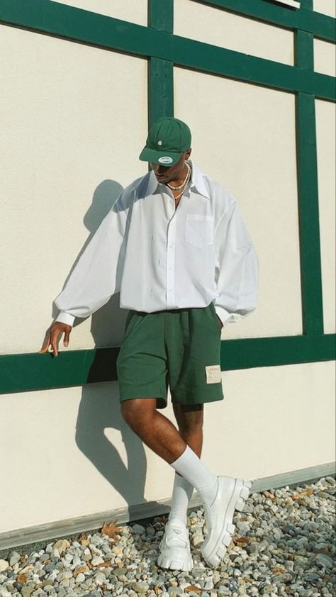 Fashionable man in white shirt and green shorts leans stylishly against a wall, wearing a cap and white sneakers.