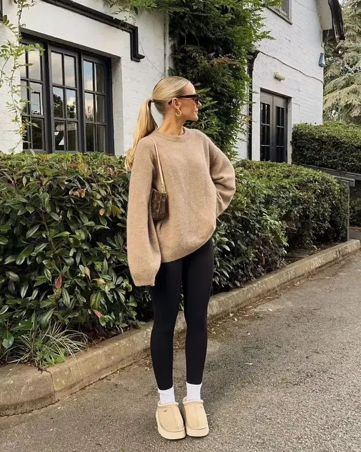 Stylish woman in cozy sweater and leggings with sunglasses standing outdoors by house and greenery.