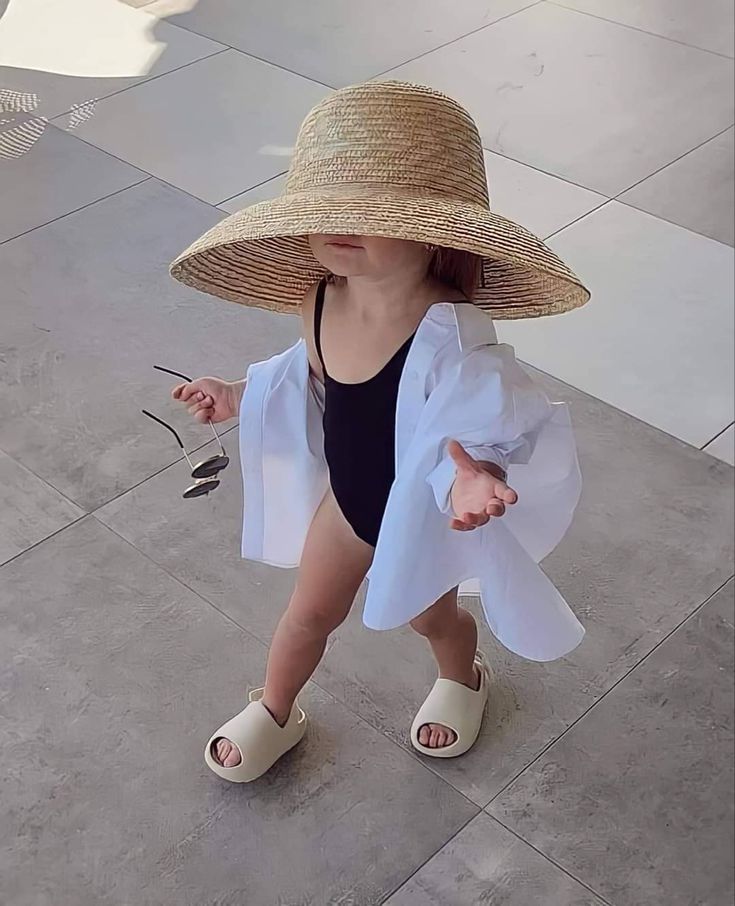 A toddler in a large sun hat, black swimsuit, white shirt, and sandals holds sunglasses on a sunny day.