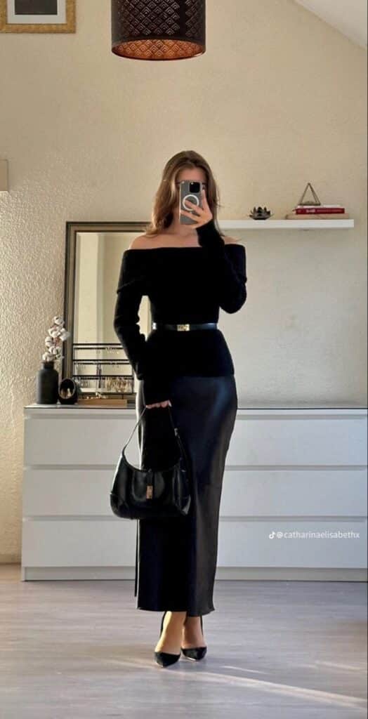 Woman in elegant black outfit takes mirror selfie, featuring off-shoulder top and skirt with belt, holding a black handbag.