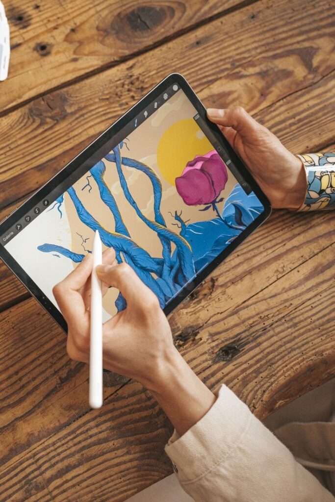 Digital artwork of a pink flower and blue vines on a tablet screen with stylus on wooden table.