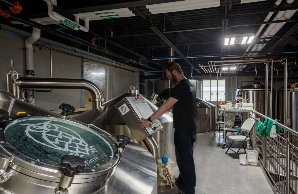 880032e6cfd9d17753eb12a55fb79f01 Man operating brewing equipment in a modern brewery, surrounded by stainless steel tanks and pipes. | Sky Rye Design Man operating brewing equipment in a modern brewery, surrounded by stainless steel tanks and pipes.