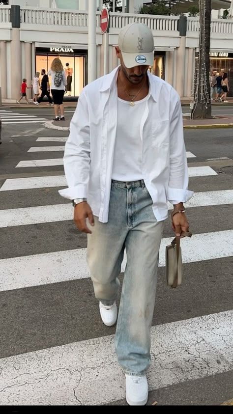 Man in casual streetwear crossing street, wearing white shirt, cap, jeans, sneakers, holding a small bag.