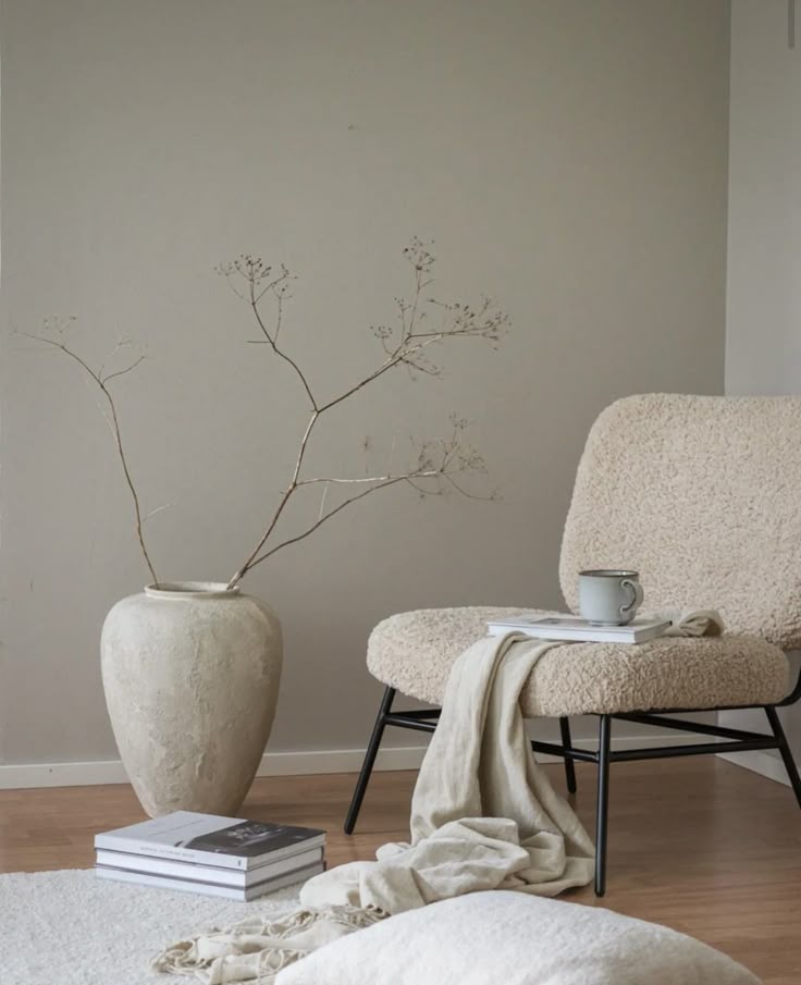 Cozy minimalist room with beige chair, vase with branches, books, and neutral accents on wooden floor.