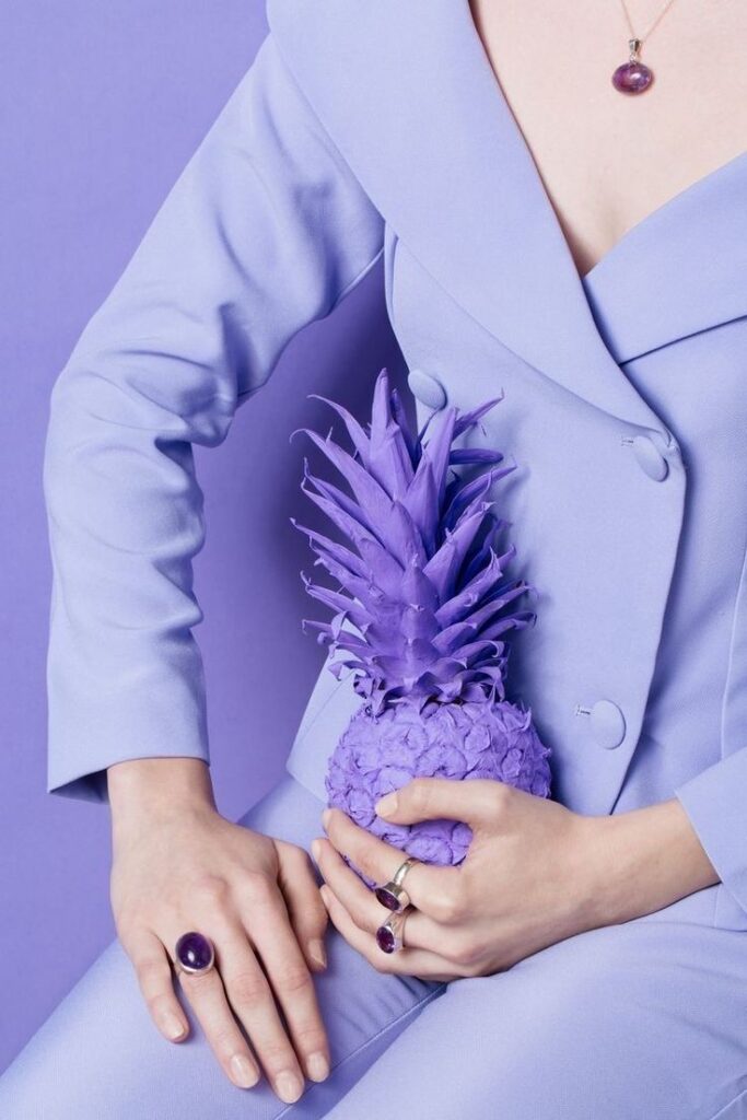 Person in lavender suit holding a purple pineapple, wearing amethyst jewelry. Vibrant monochromatic aesthetic.