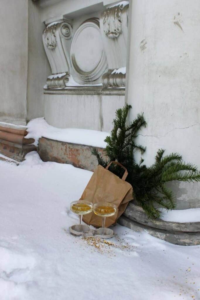 Two champagne glasses and a paper bag with greenery on snowy ledge of ornate building.