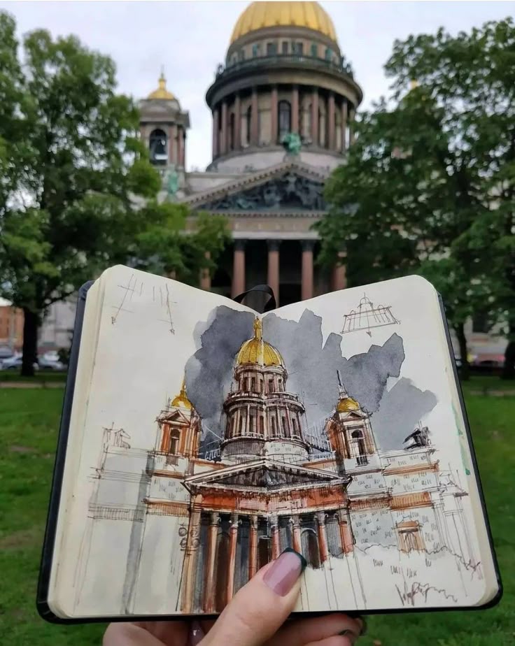 Sketchbook with drawing of cathedral dome held up against actual building in a park setting.