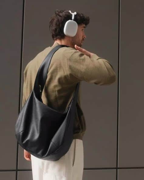 Man wearing headphones with a black leather bag stands against a modern wall, looking away in a stylish, casual outfit.