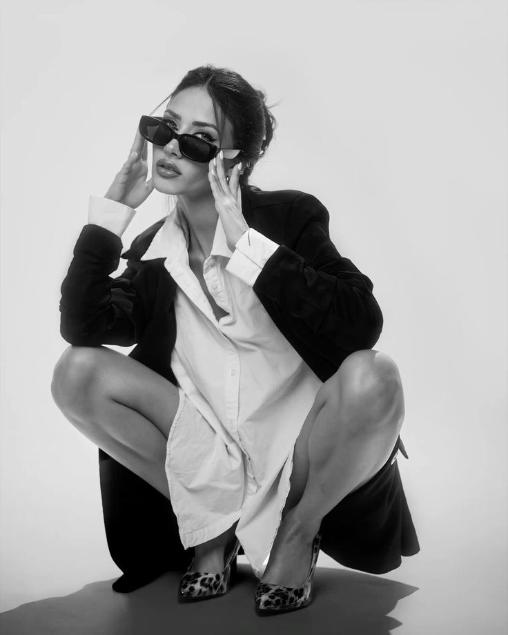 Stylish woman in sunglasses squatting, wearing a chic outfit with leopard print heels and a loose shirt. Black and white photo.