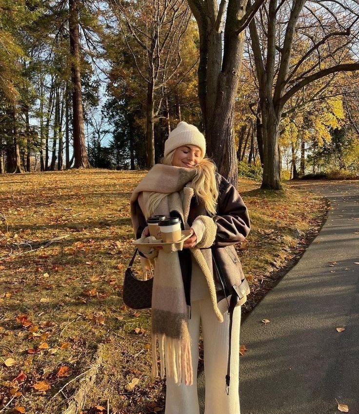 Woman enjoying autumn walk in park, bundled in scarf and hat, carrying coffee. Sunlit trees with colorful leaves surround her.