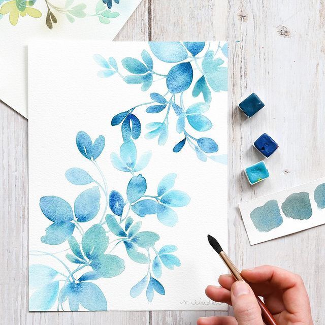 Watercolor painting of blue leaves on paper with paintbrush and blue paint.