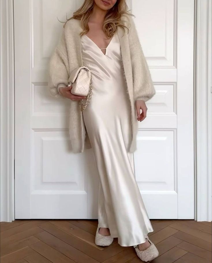 Woman in elegant white satin dress with knit cardigan and matching purse, standing in a stylish room.