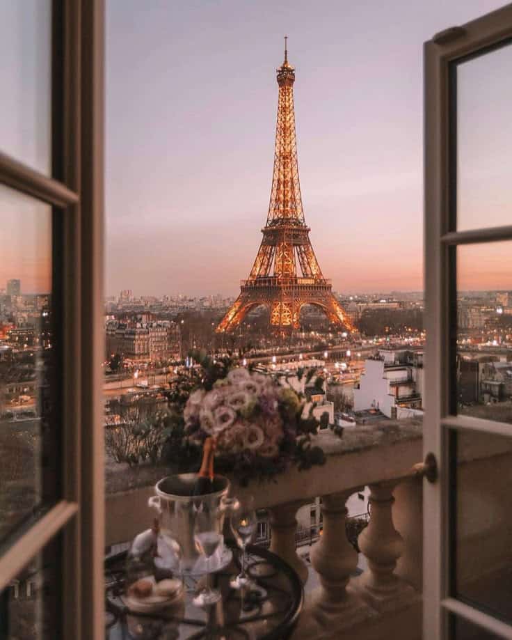 View of illuminated Eiffel Tower at sunset from a balcony, with flowers and champagne, Paris romance scene.