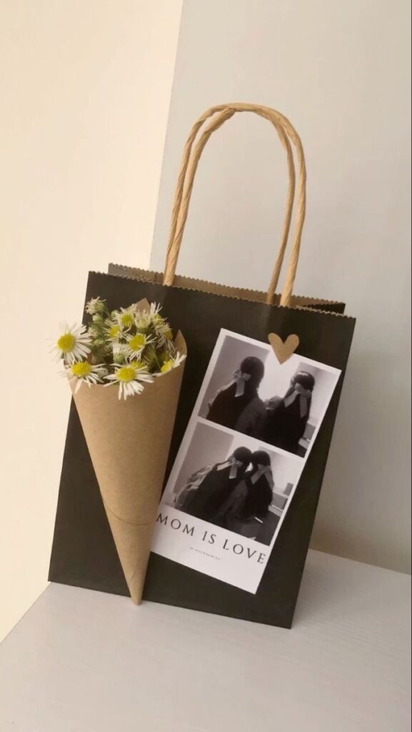 947c4115-3d2c-4239-80d3-07fe65db4418 Gift bag with daisy bouquet and photo collage, Mom is Love message on table. | Sky Rye Design Gift bag with daisy bouquet and photo collage, Mom is Love message on table.