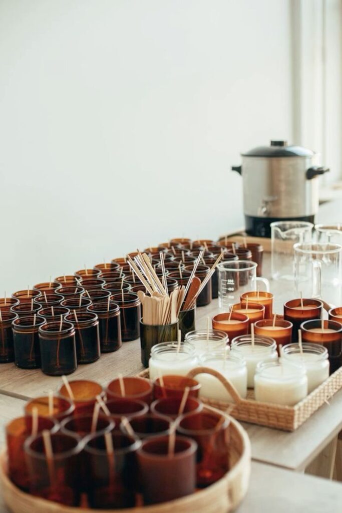 Handmade candles in jars with wax melting equipment on a wooden table, ready for crafting and packaging.