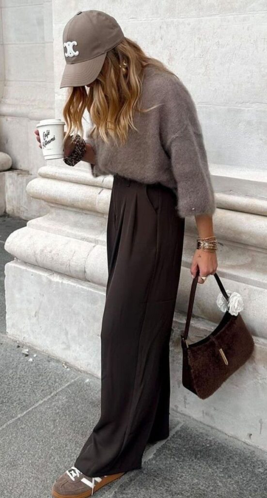 Fashionable woman in casual fall outfit with coffee cup, brown trousers, and stylish cap, holding a chic handbag.