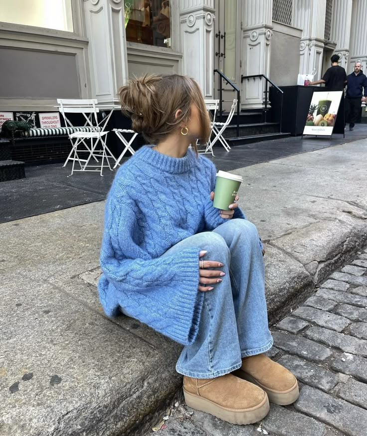 Woman in oversized blue sweater sits on sidewalk, holds coffee cup, wearing jeans and boots, near outdoor cafe seating.