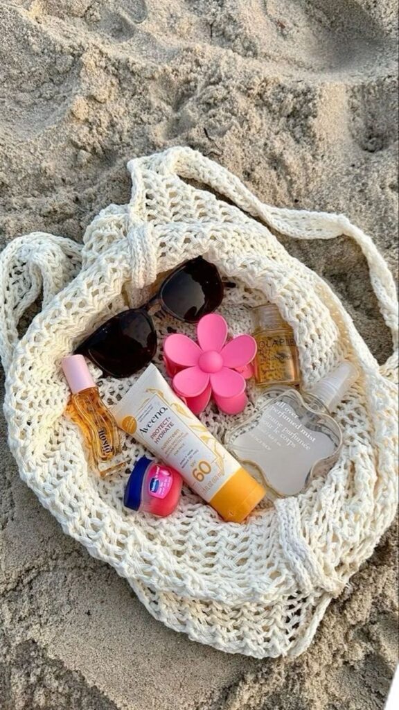 Beach essentials in woven bag: sunglasses, sunscreen, lip balm, flower accessory, on sandy shore.
