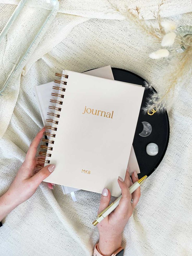 Person holding a journal and pen on a cozy beige blanket, with decorative crystals and flowers nearby.