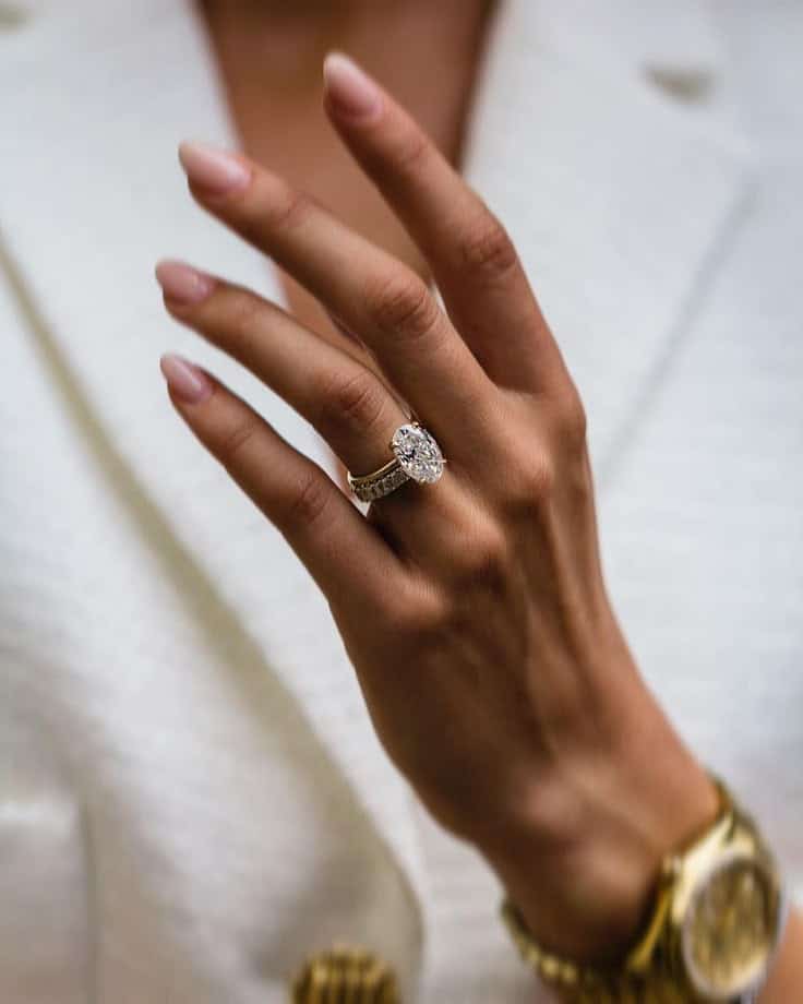 Elegant hand showcasing an oval diamond ring on a gold band, paired with a stylish watch against a white blazer backdrop.