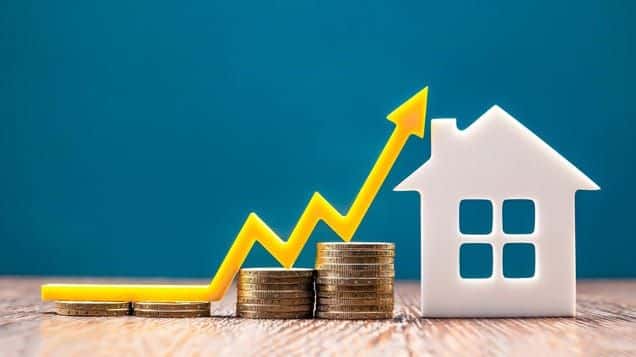 Rising home prices concept with stacked coins, arrow graph, and house icon on wooden surface against blue background.