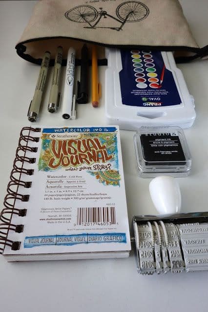 Art supplies including pens, watercolor set, visual journal, and stamp are arranged on a desk.
