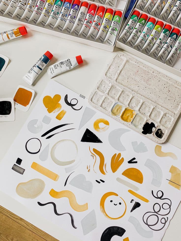 Art supplies and a palette with colorful gouache paintings on paper, showcasing abstract designs and shapes.