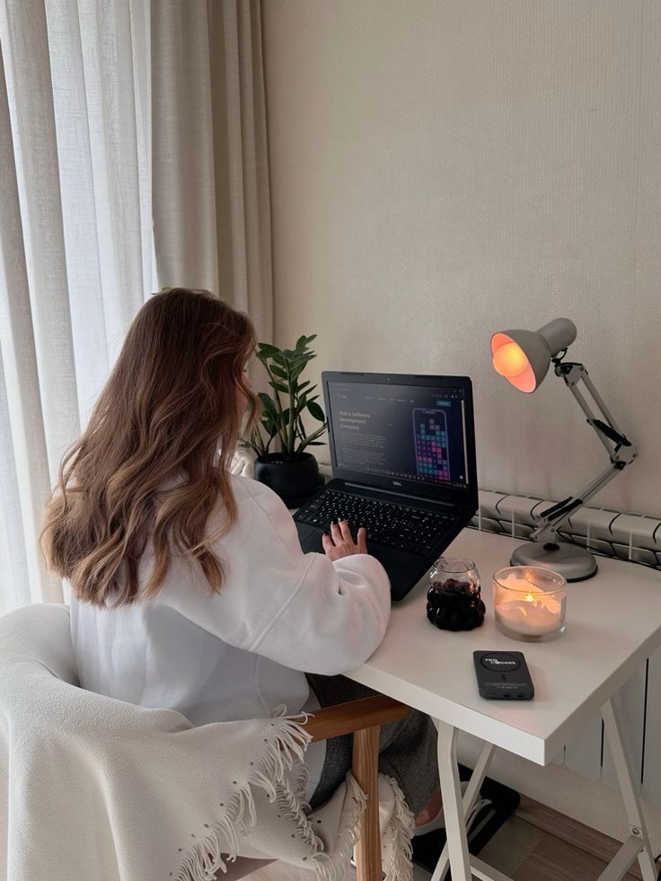 Woman working on a laptop at a home desk with a cozy setup, featuring candles and a lamp.