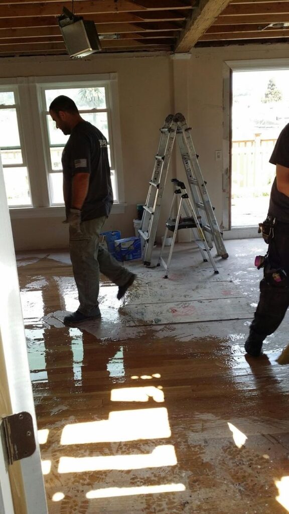Construction workers renovating room with exposed ceiling, ladders, and wet floor.