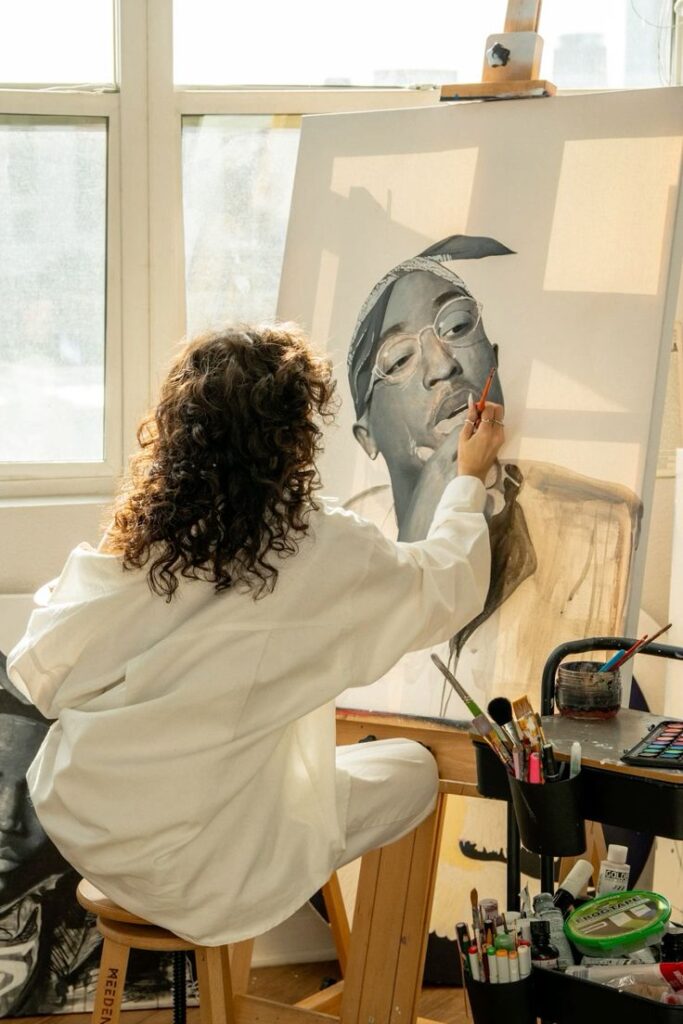 Artist painting a portrait on canvas in a sunlit studio, surrounded by art supplies.