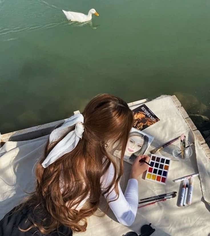 Woman painting by a lakeside, with art supplies and a duck swimming nearby.