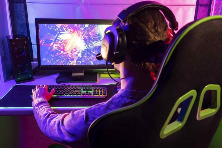 Gamer with headset playing a colorful video game on a PC with RGB lighting, sitting in a gaming chair.