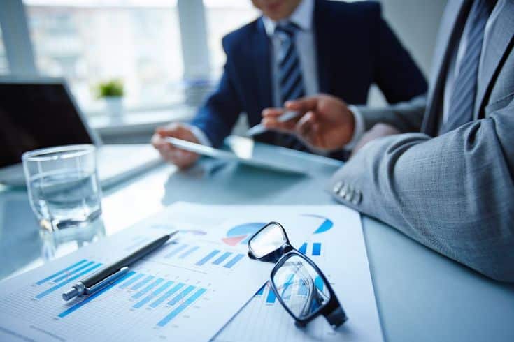Two business professionals discussing charts at a desk with reports, glasses, and a pen in focus.
