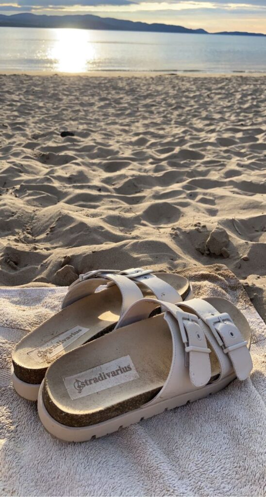 White sandals on a sandy beach during sunset, ocean and distant mountains in the background.