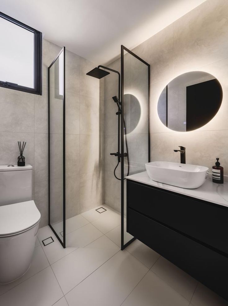 Modern bathroom with glass shower, round mirror, vanity, and toilet, featuring elegant black and white decor.