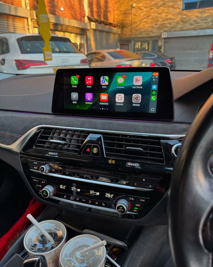 Car interior with dashboard display showing apps, climate control, and two drinks in holder.