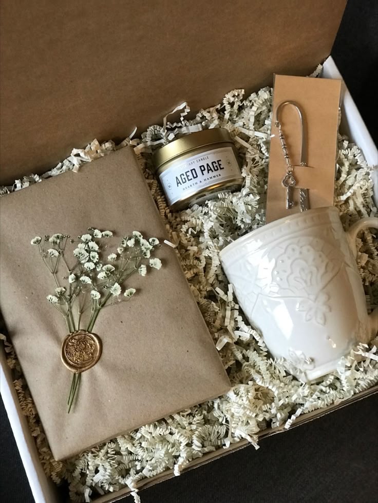 Gift box with a candle, white mug, notebook with dried flowers, and a silver bookmark, nestled in decorative filler.