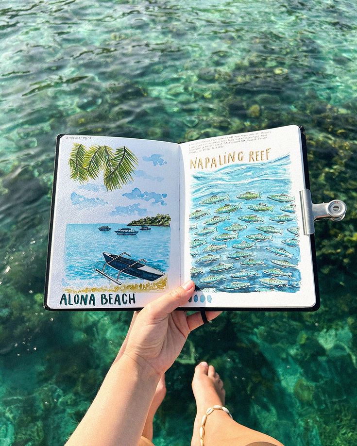 Hand holding an open sketchbook with Alona Beach and Napaling Reef illustrations over clear turquoise water.
